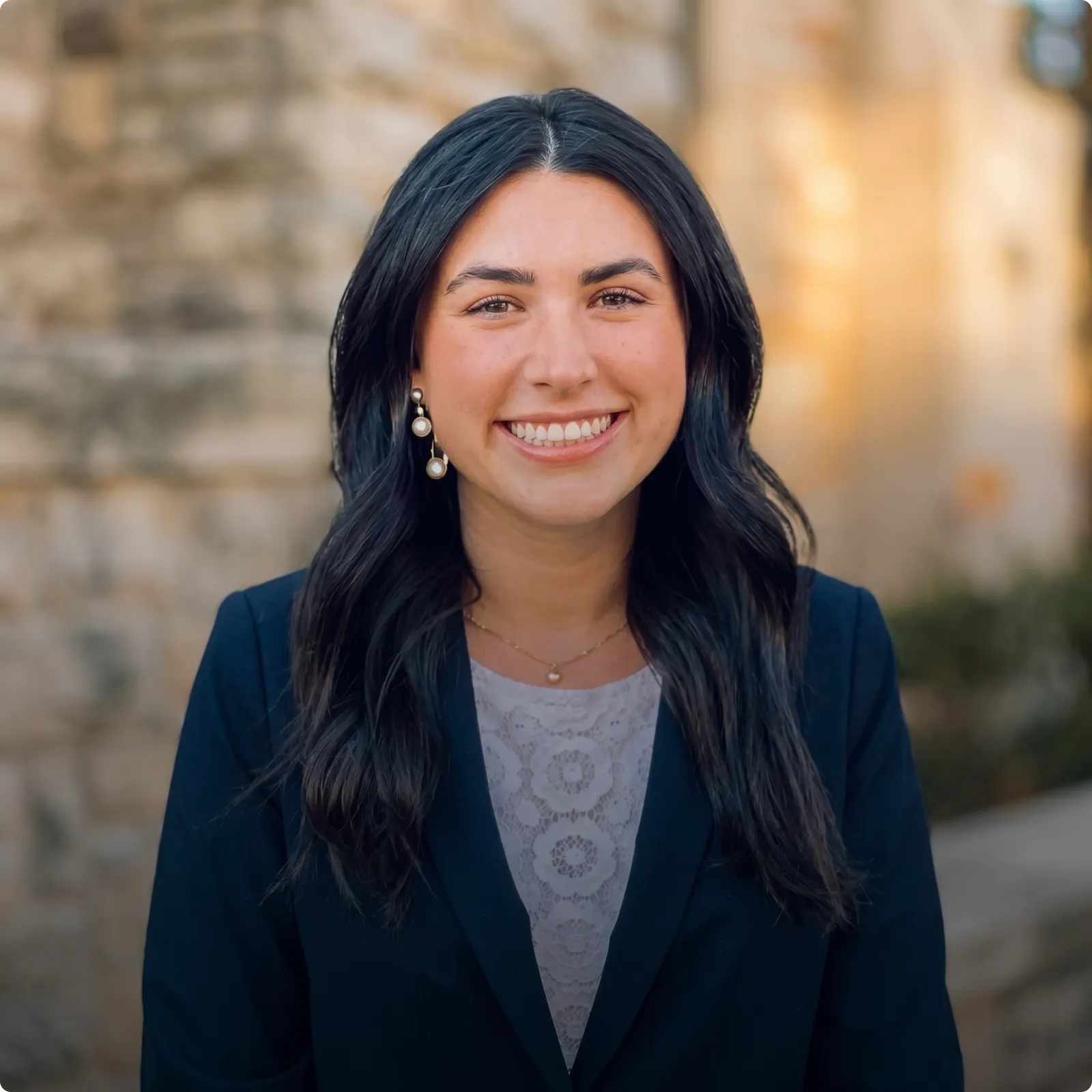 HOA attorney / HOA law A woman with long dark hair and a professional smile, wearing a white lace top and a navy blazer, in front of a blurred background.