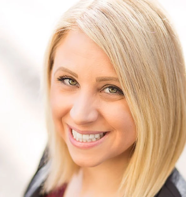HOA attorney / HOA law A woman with blonde hair and a friendly smile, looking directly at the camera. She is wearing a black and white plaid shirt and has a subtle, natural makeup look. The background is a soft, white or light gray color, and the overall atmosphere is professional and friendly.
